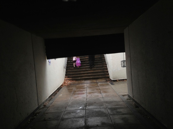 Looking out of the tunnel we see a little girl dressed as a princess from behind, climbing the stairs in the rain