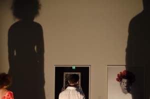 The woman and man's shadows are tall against the wall and cast across images on the gallery wall of a man and mirror with water in its frame
